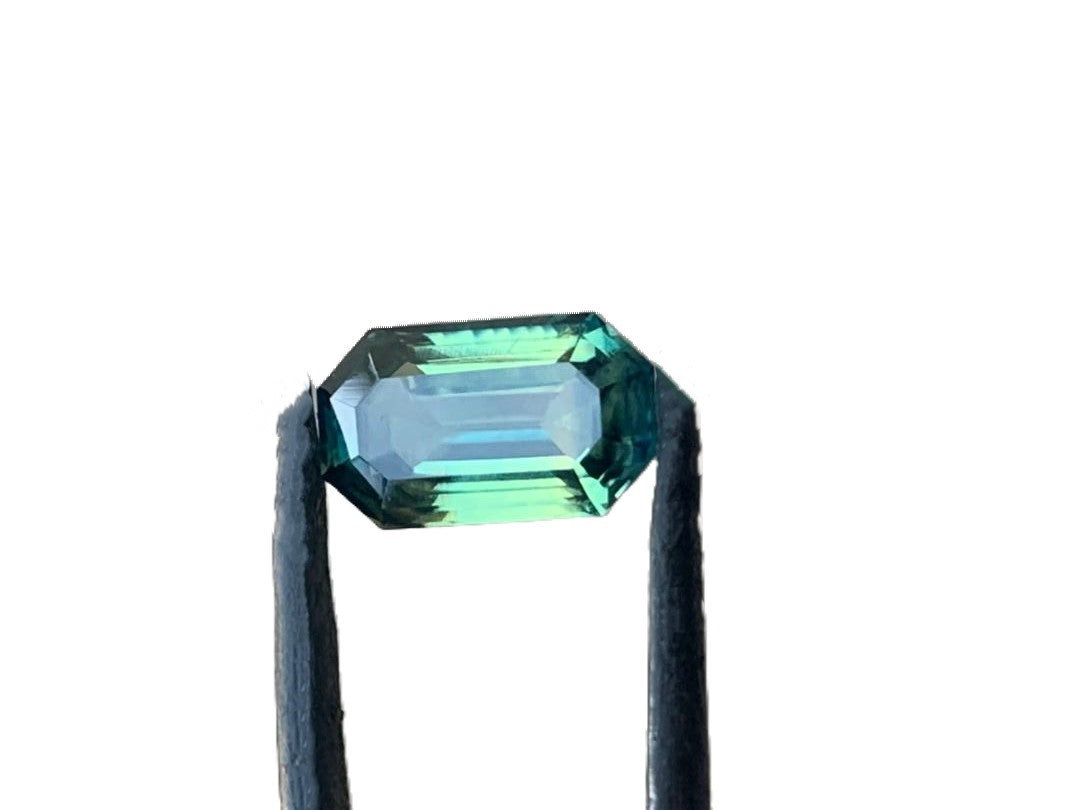 Loupe clean emerald cut teal blue sapphire gemstone held by tweezers against a white background, emphasizing its vibrant blue-green tones.