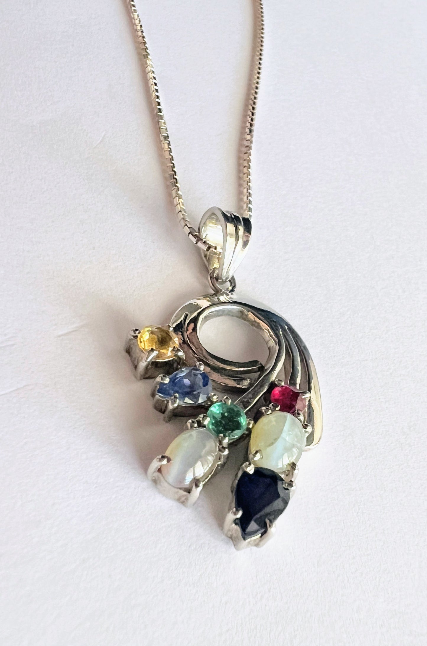 Angled view of Multigem Swirling Infinity Pendant Necklace highlighting colorful gemstones in floral motif. Features ruby, emerald, and natural sapphires.