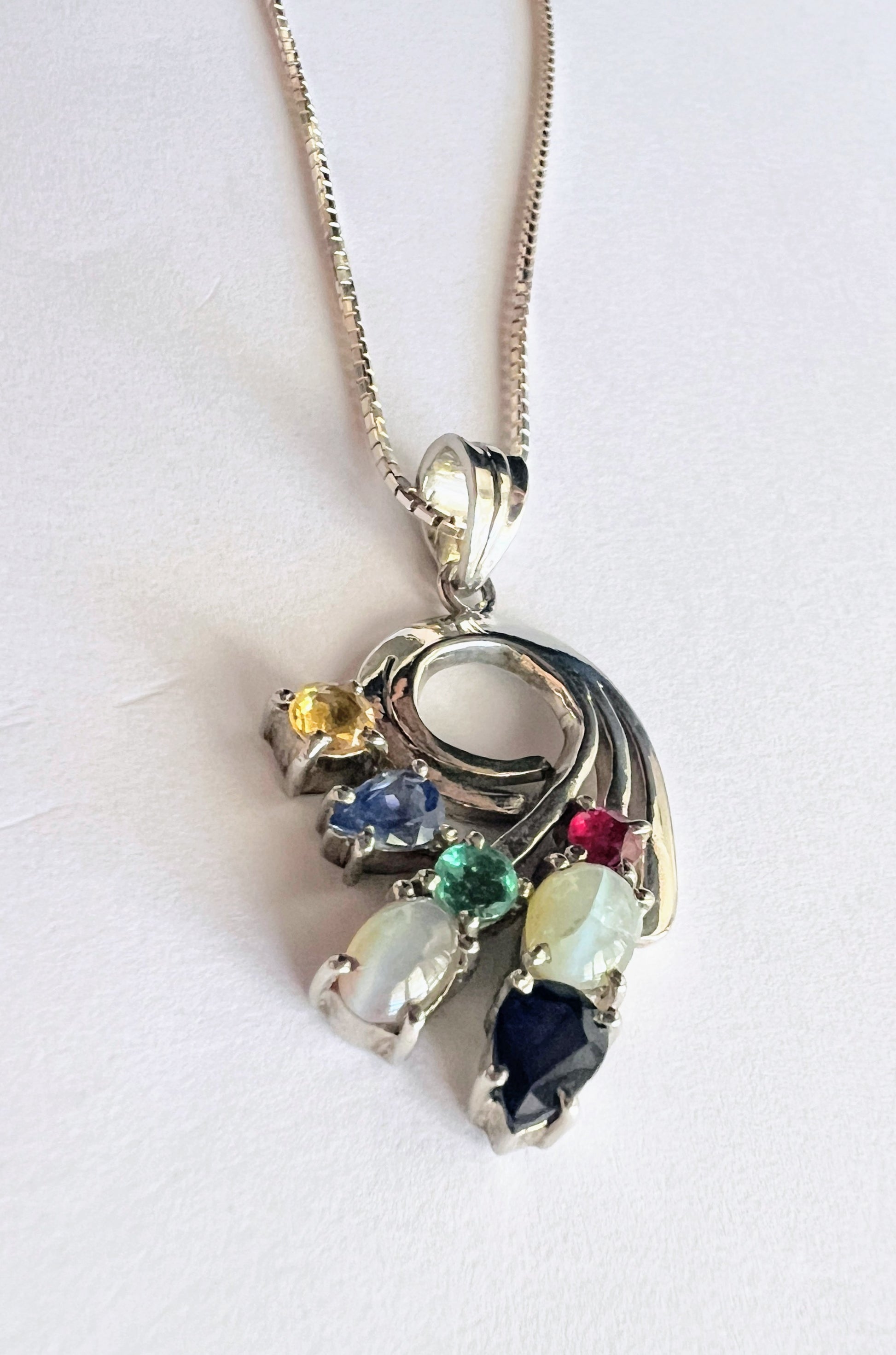 Angled view of Multigem Swirling Infinity Pendant Necklace highlighting colorful gemstones in floral motif. Features ruby, emerald, and natural sapphires.