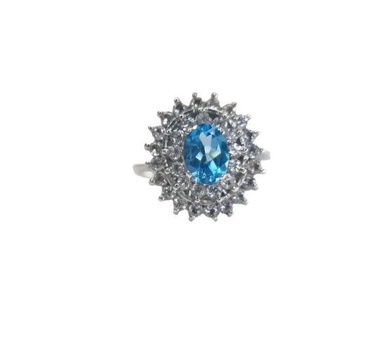 A 2ct Oval Blue Topaz Silver Cocktail Ring with a double halo of white zircons, placed on a white surface.