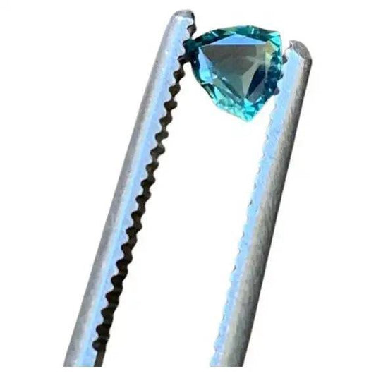 0.5ct trillion cut teal blue sapphire gemstone held by metal tweezers, showcasing its brilliant facets and deep blue-green hue.