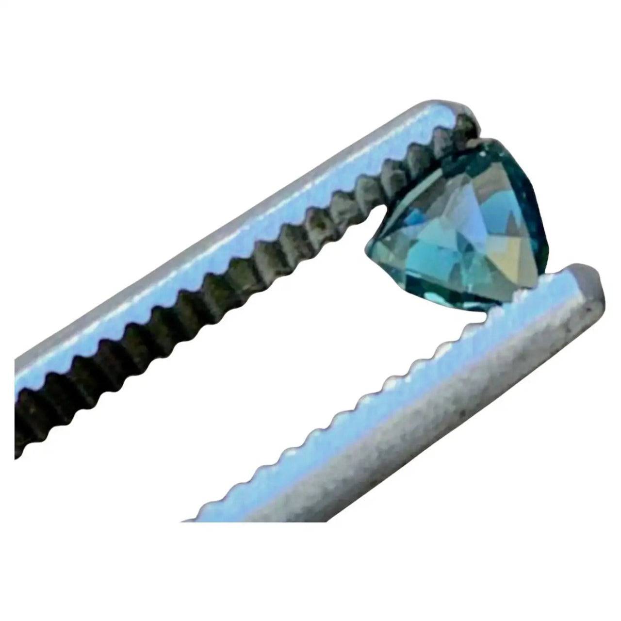 Close-up of a 0.5ct trillion cut teal blue sapphire gemstone, held by tweezers, highlighting its clarity and vibrant color.