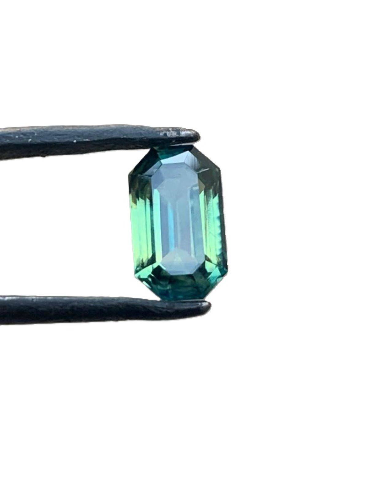 Loupe clean 1.8ct emerald cut teal blue sapphire gemstone held by tweezers against a white background, reflecting its deep blue-green hues.
