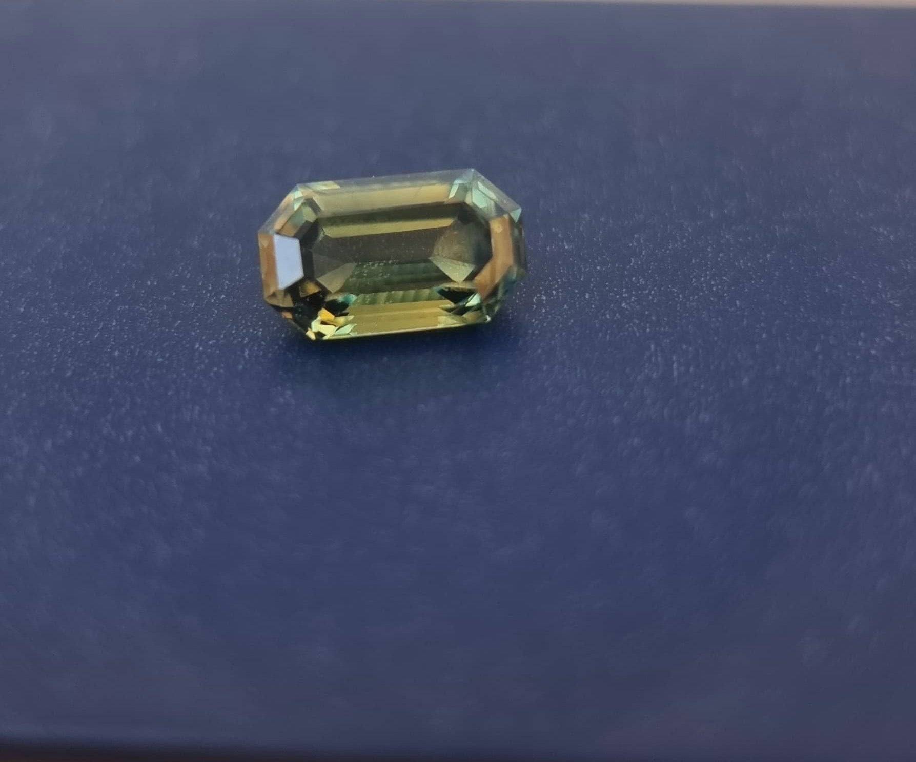 Loose teal blue sapphire gemstone with an emerald cut placed on a dark surface, highlighting its natural brilliance and faceted design.