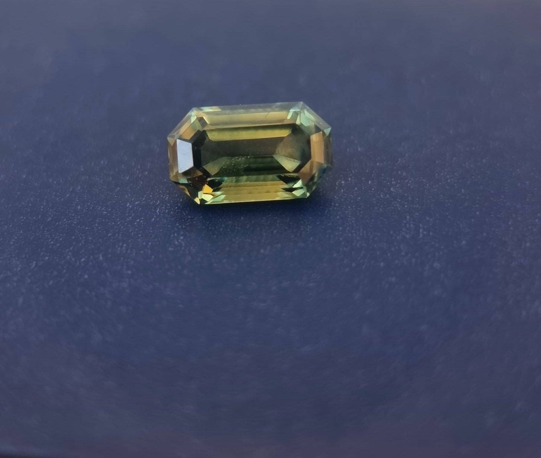 Teal blue sapphire gemstone with an emerald cut resting on a dark surface, reflecting its natural clarity and elegant faceting.