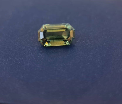 Teal blue sapphire gemstone with an emerald cut resting on a dark surface, reflecting its natural clarity and elegant faceting.