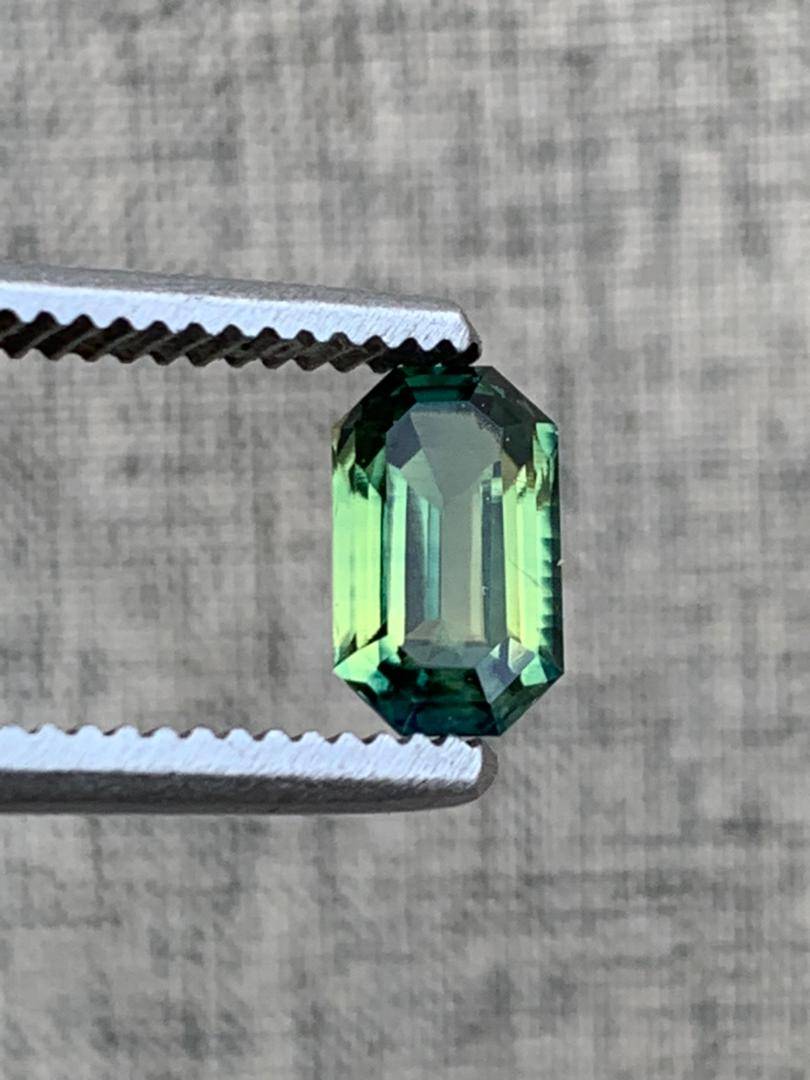 Loupe clean 1.8ct emerald cut teal blue sapphire gemstone held by metal tweezers against a textured background, showcasing its brilliance.