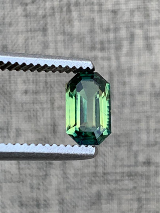 Loupe clean 1.8ct emerald cut teal blue sapphire gemstone held by metal tweezers against a textured background, showcasing its brilliance.