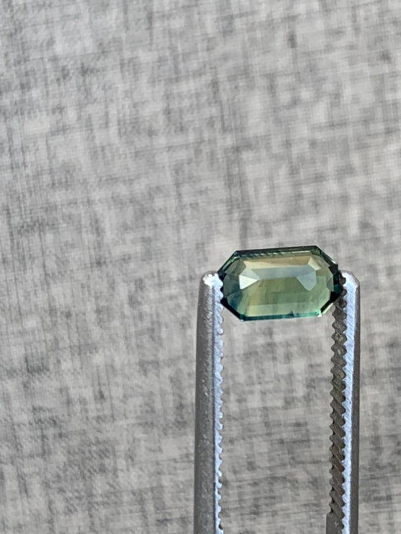 Emerald cut teal blue sapphire gemstone held by silver tweezers against a textured background, showcasing its elegant cut and transparency.