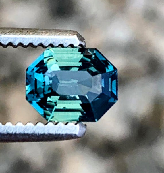 High-quality 2.5ct radiant cut teal blue sapphire gemstone held by tweezers, showcasing its deep blue-green hues and precision cut.